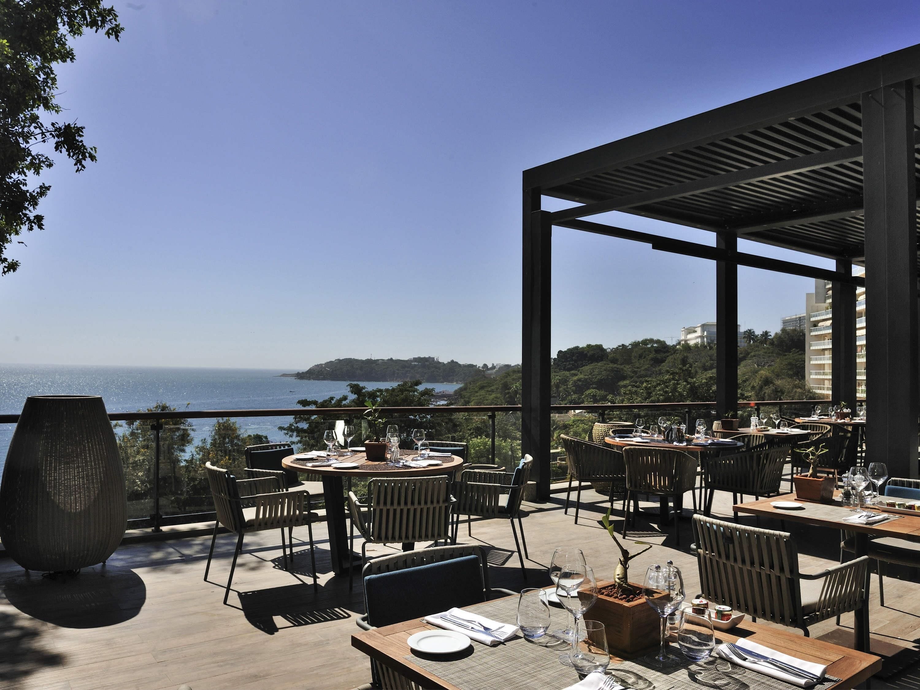Un área de comedor al aire libre con mesas y sillas, con vistas panorámicas al mar y las colinas.