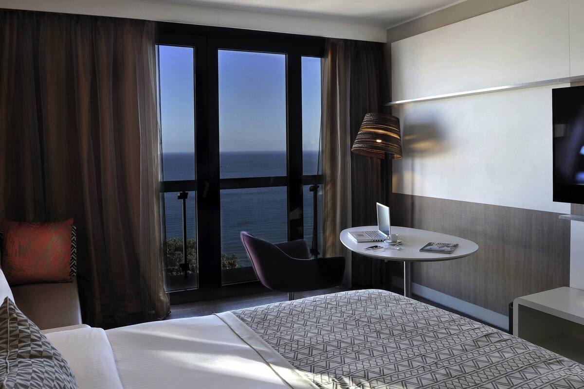 A hotel room with a bed, a desk with a laptop, a chair, a TV, and a view of the ocean.