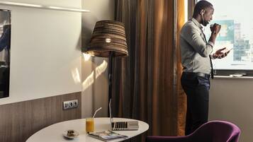 Habitación de hotel con una laptop sobre una mesa, una lámpara, una silla morada y una ventana que ofrece vistas a la ciudad.
