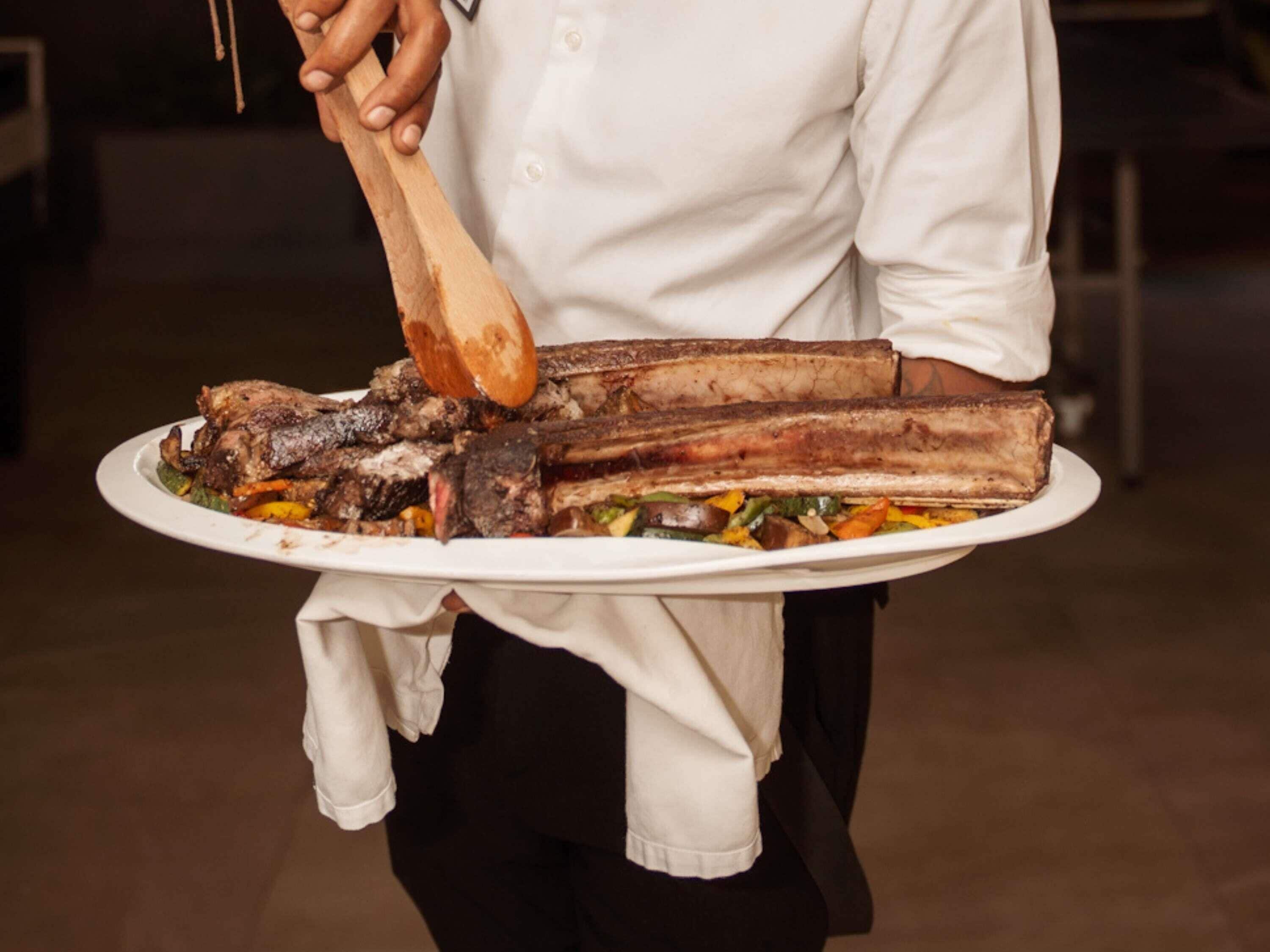 A person holding a large white plate with a roasted pig leg and vegetables.