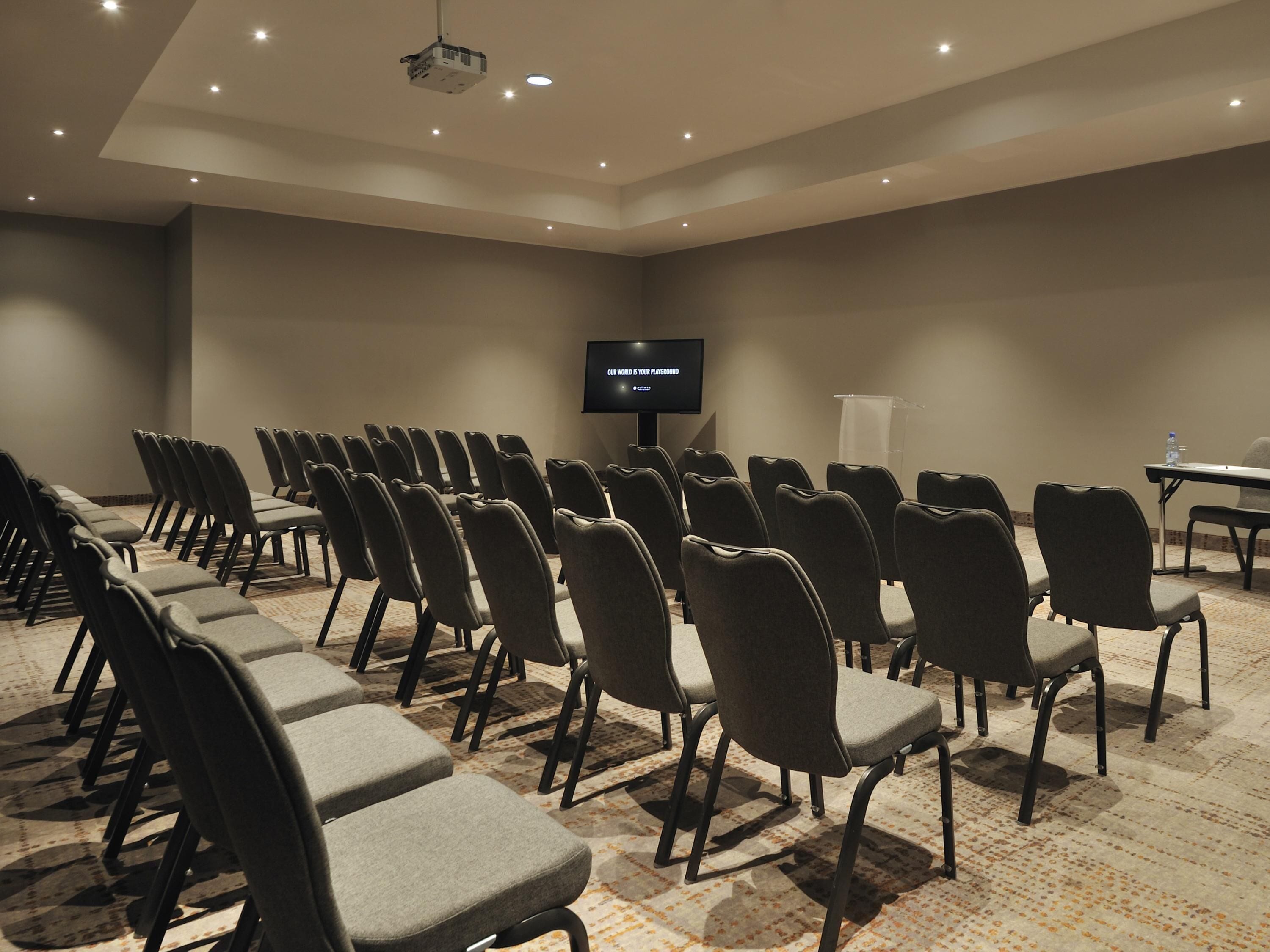 A conference room with rows of chairs, a podium, a projector, and a screen displaying 'OUR WORLD IS YOUR PLATFORM'.