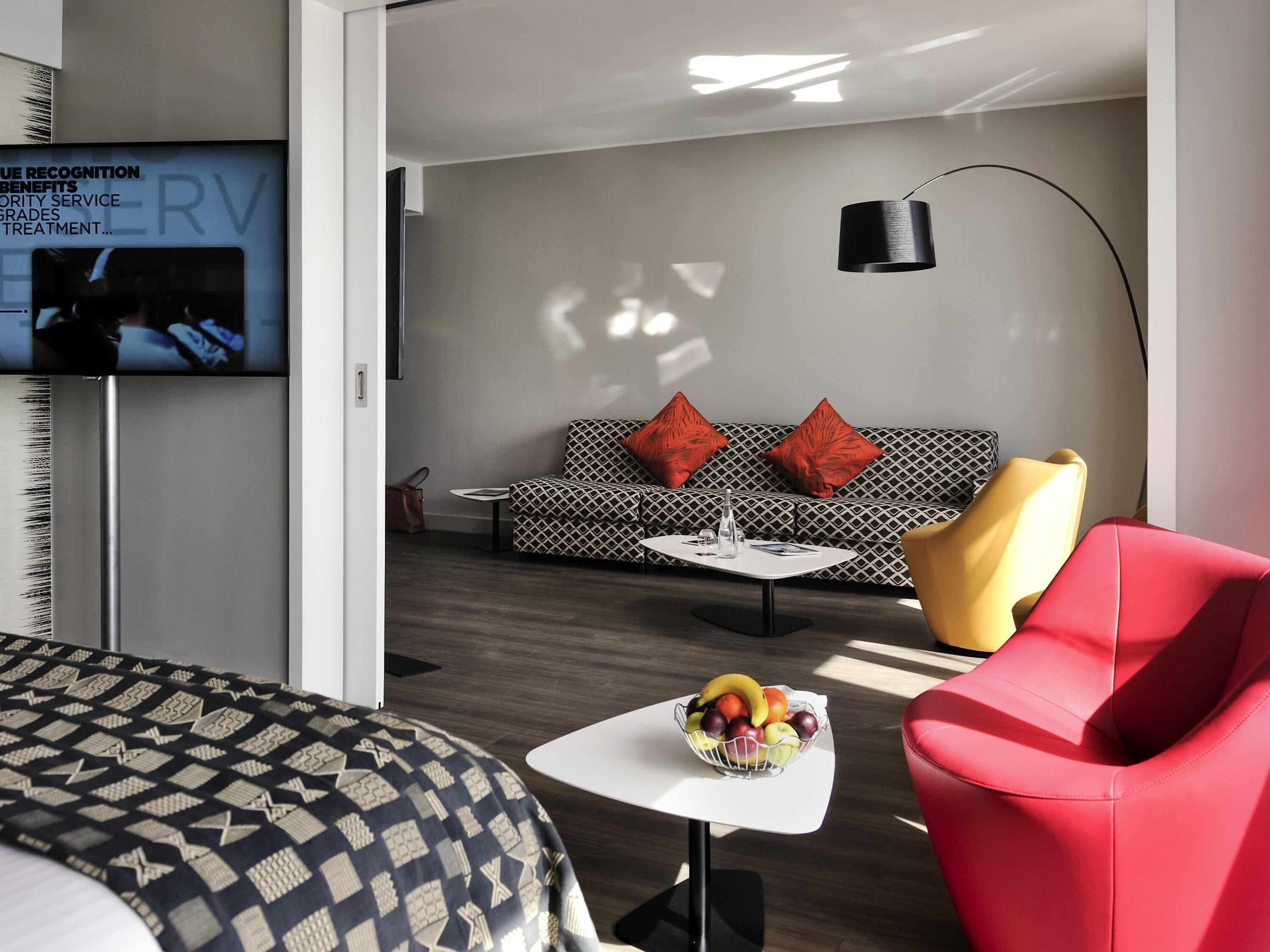Ein modernes Hotelzimmer mit einer Couch, einem roten Sessel, einem kleinen Tisch mit Obstschale und einem an der Wand befestigten Fernseher.