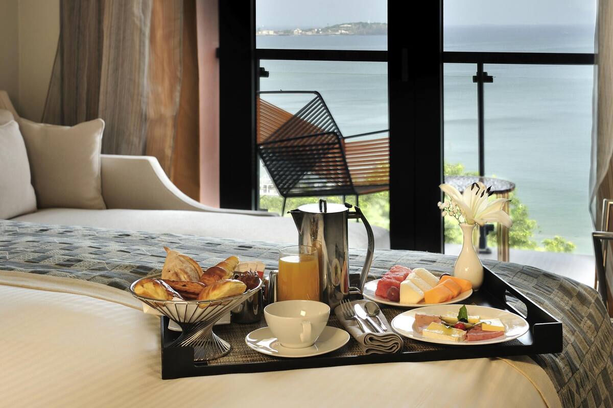 A bed with a tray of breakfast items, a vase of flowers, and a view of the sea.