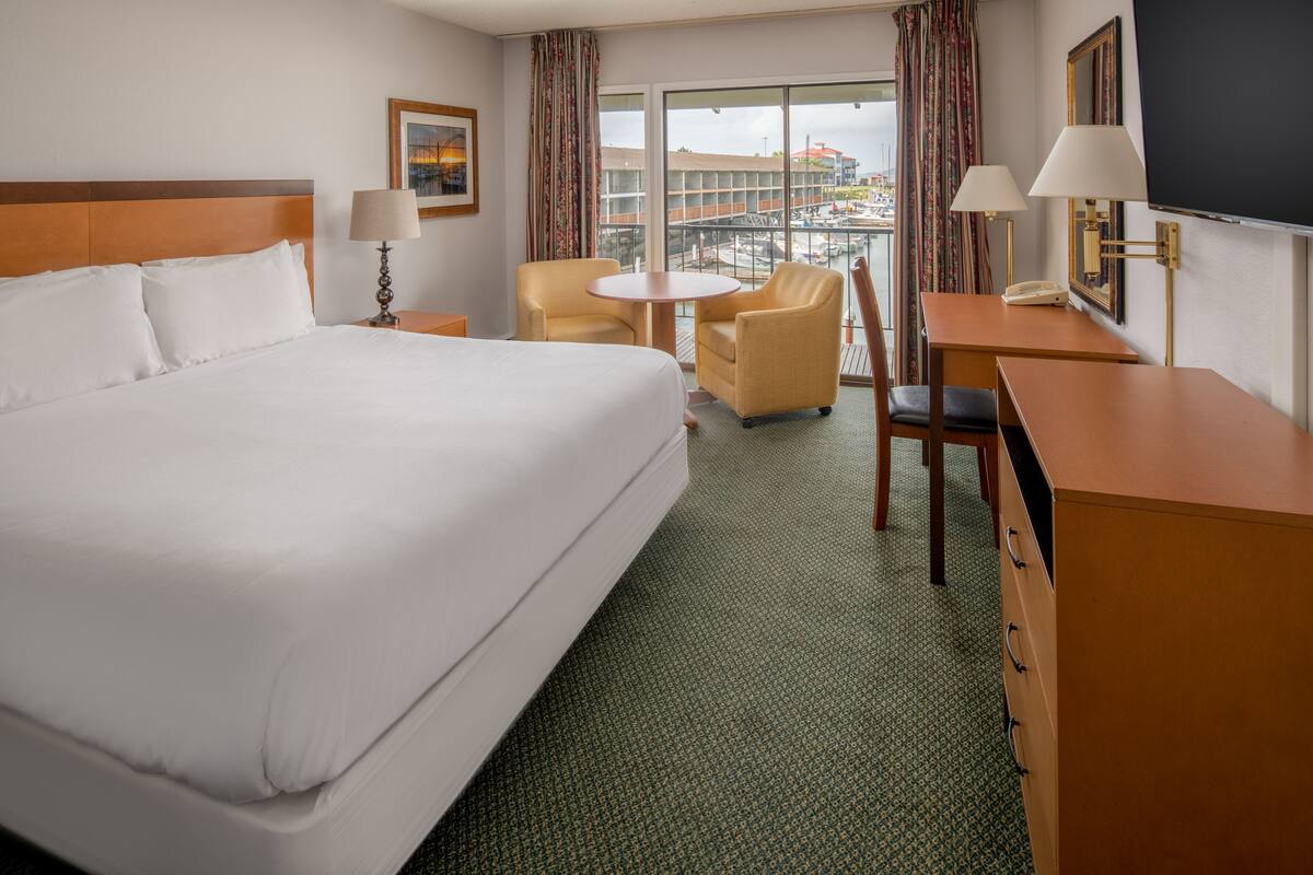 Habitación de hotel con una cama grande, un escritorio, dos sillas, una mesita y vistas a la ciudad a través de amplios ventanales.