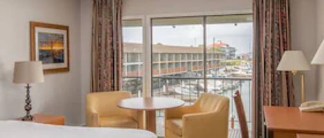 A hotel room with a bed, two armchairs, a small table, and a view of buildings through large windows.