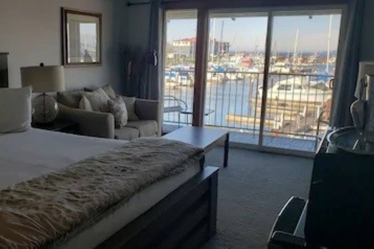 Habitación de hotel con una cama grande, un sofá, un escritorio y un balcón con vistas al puerto deportivo.