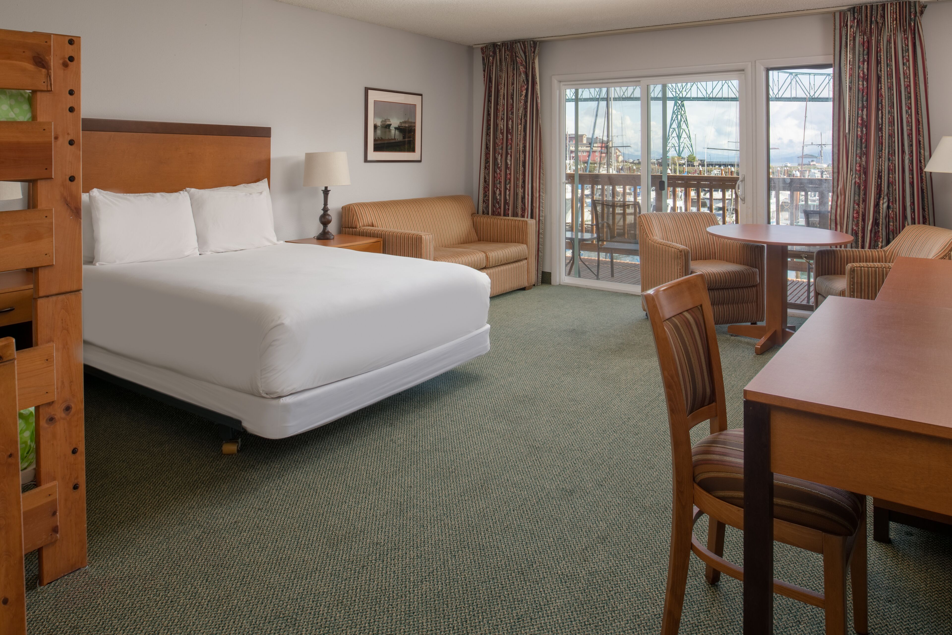 Habitación de hotel con una cama grande, una mesa de comedor y un balcón con vista a un puente y al paisaje urbano.