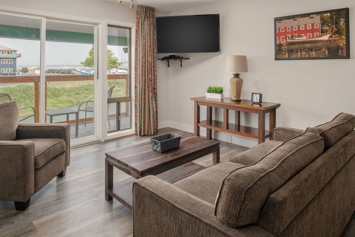 Sala de estar con sofá, sillón y una mesa de centro. Hay un televisor montado en la pared, una lámpara y un marco de fotos.