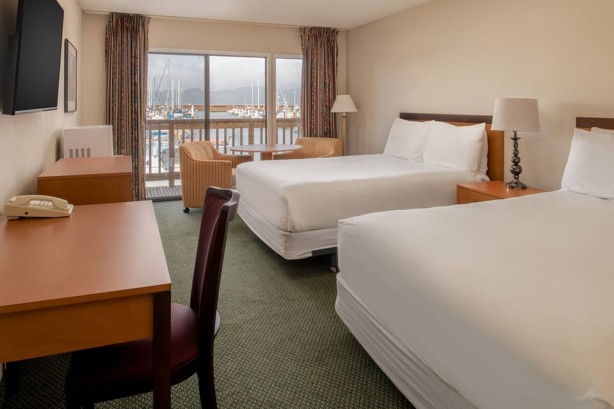 Habitación de hotel con dos camas, un escritorio, una silla, una mesa y vistas al puerto deportivo.