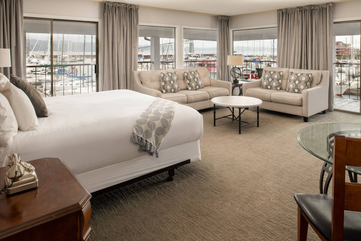 Habitación de hotel con una cama grande, un sofá, una mesa redonda y vistas a un puerto deportivo a través de amplios ventanales.