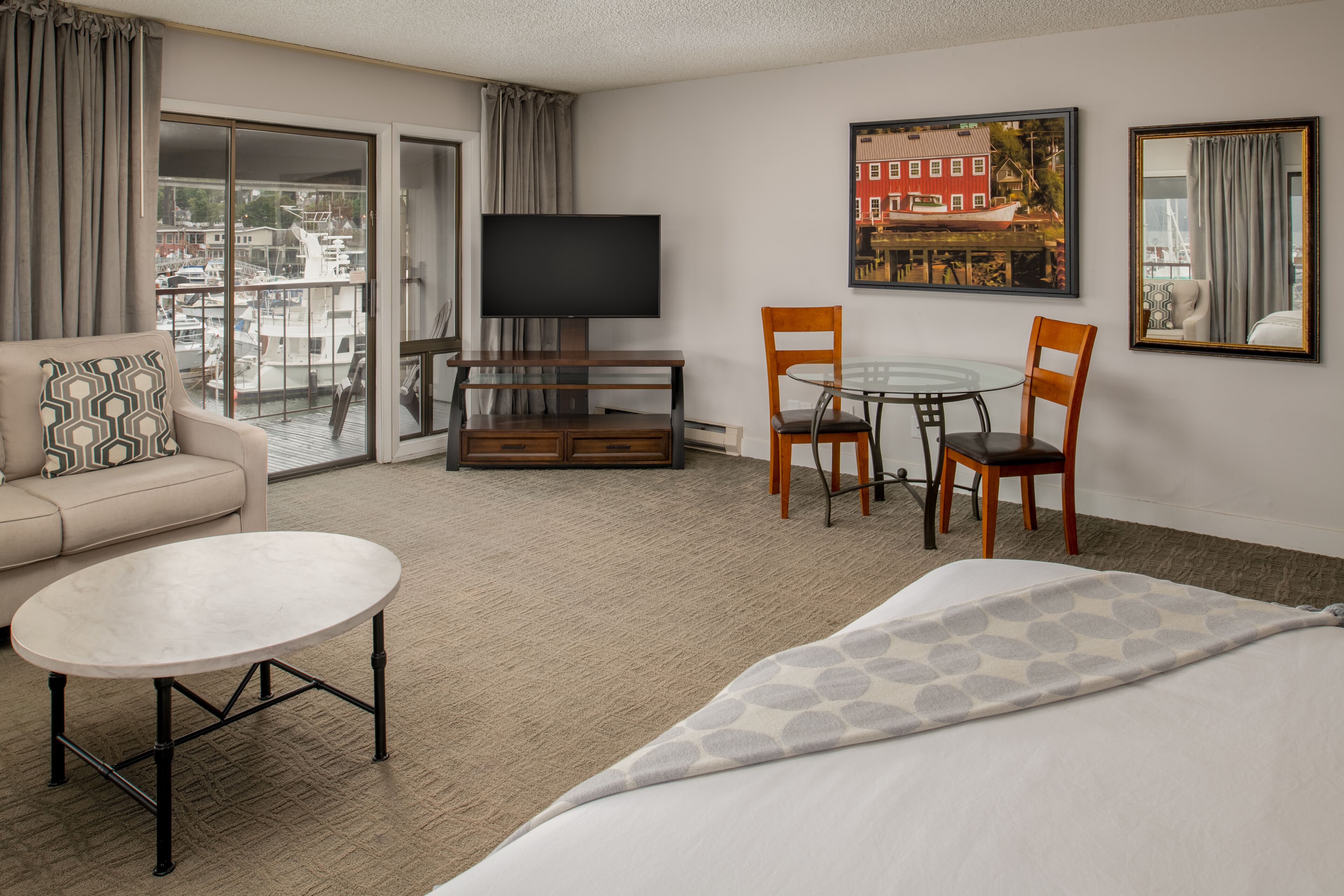 Una camera d'albergo con un letto, un divano, una TV, un tavolo da pranzo con sedie e un'ampia finestra vista barche ed edifici.