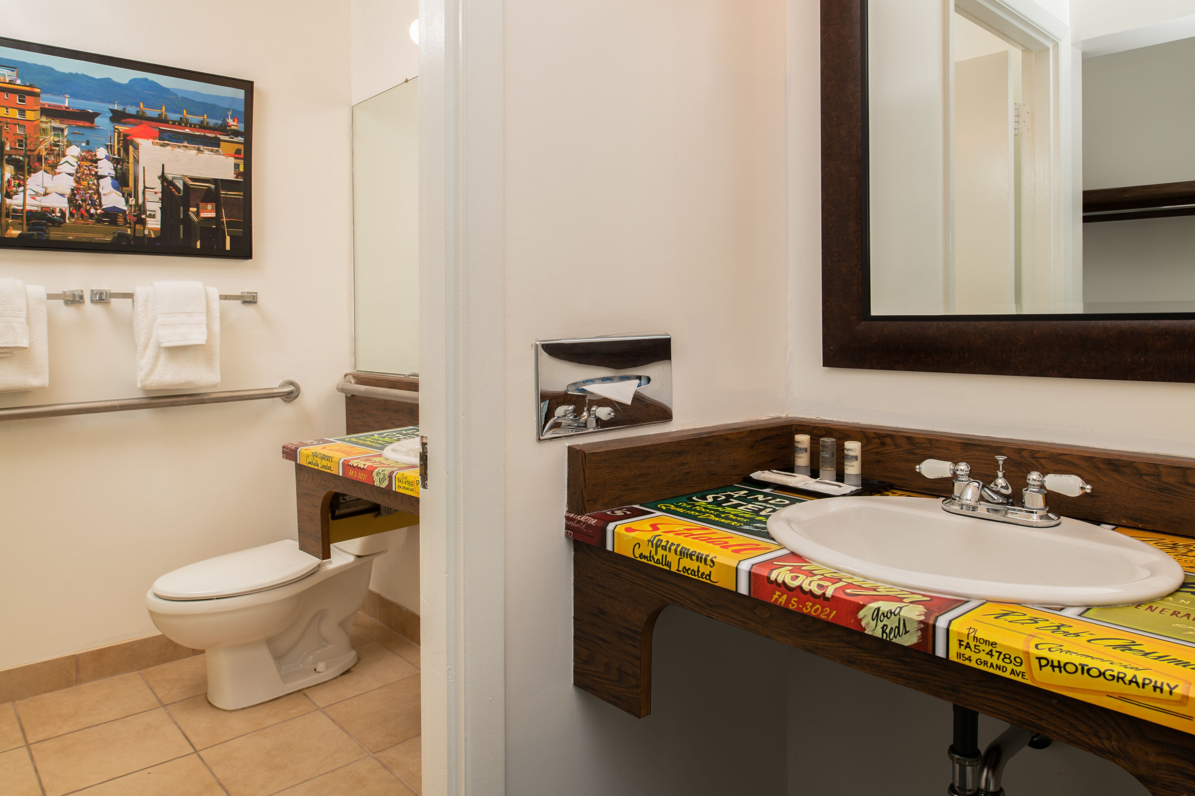 A bathroom with a toilet, a sink, and a mirror. There is a towel rack with towels and a framed picture on the wall.