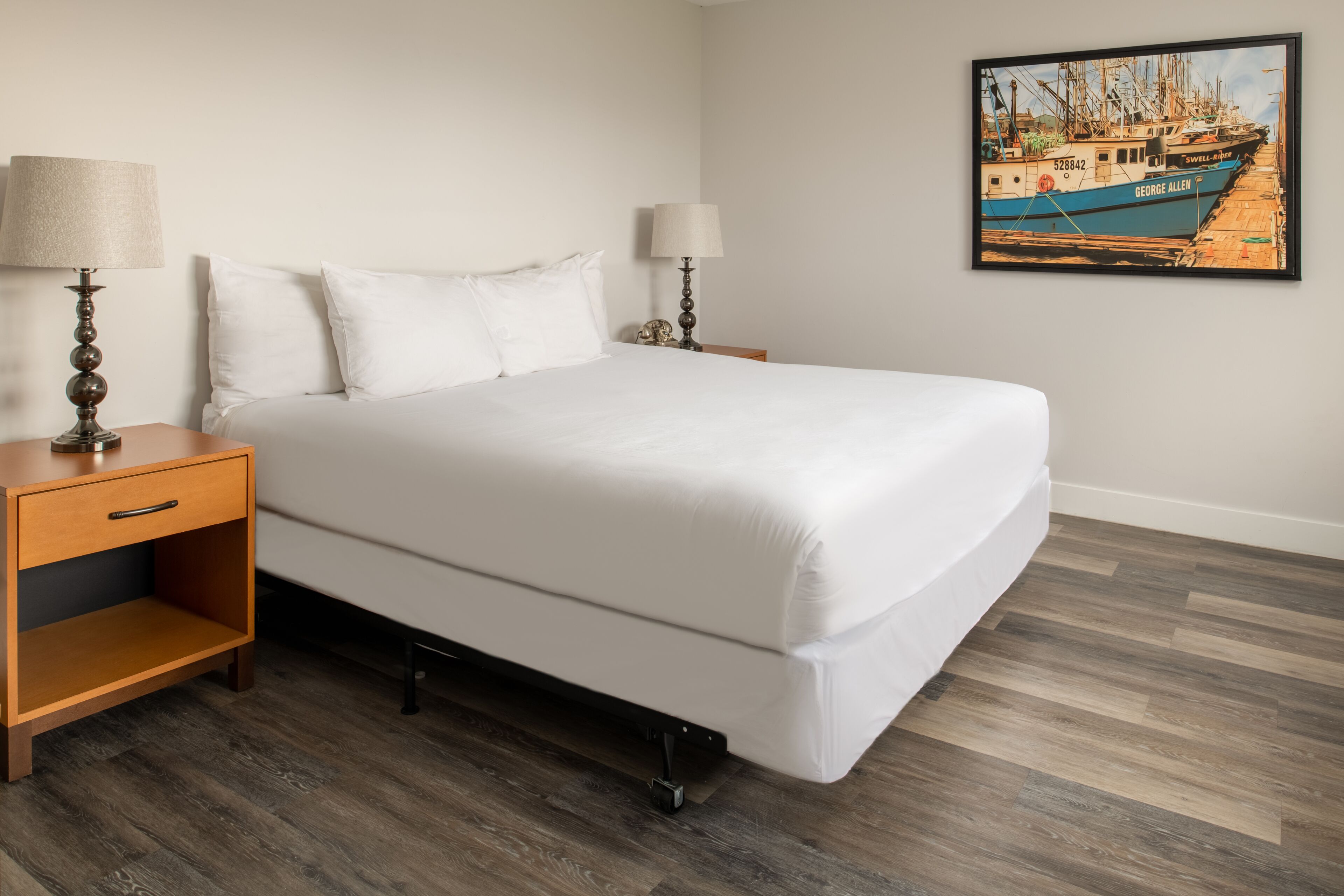 A neatly made bed with white linens, two bedside tables with lamps, and a framed picture of boats on the wall.