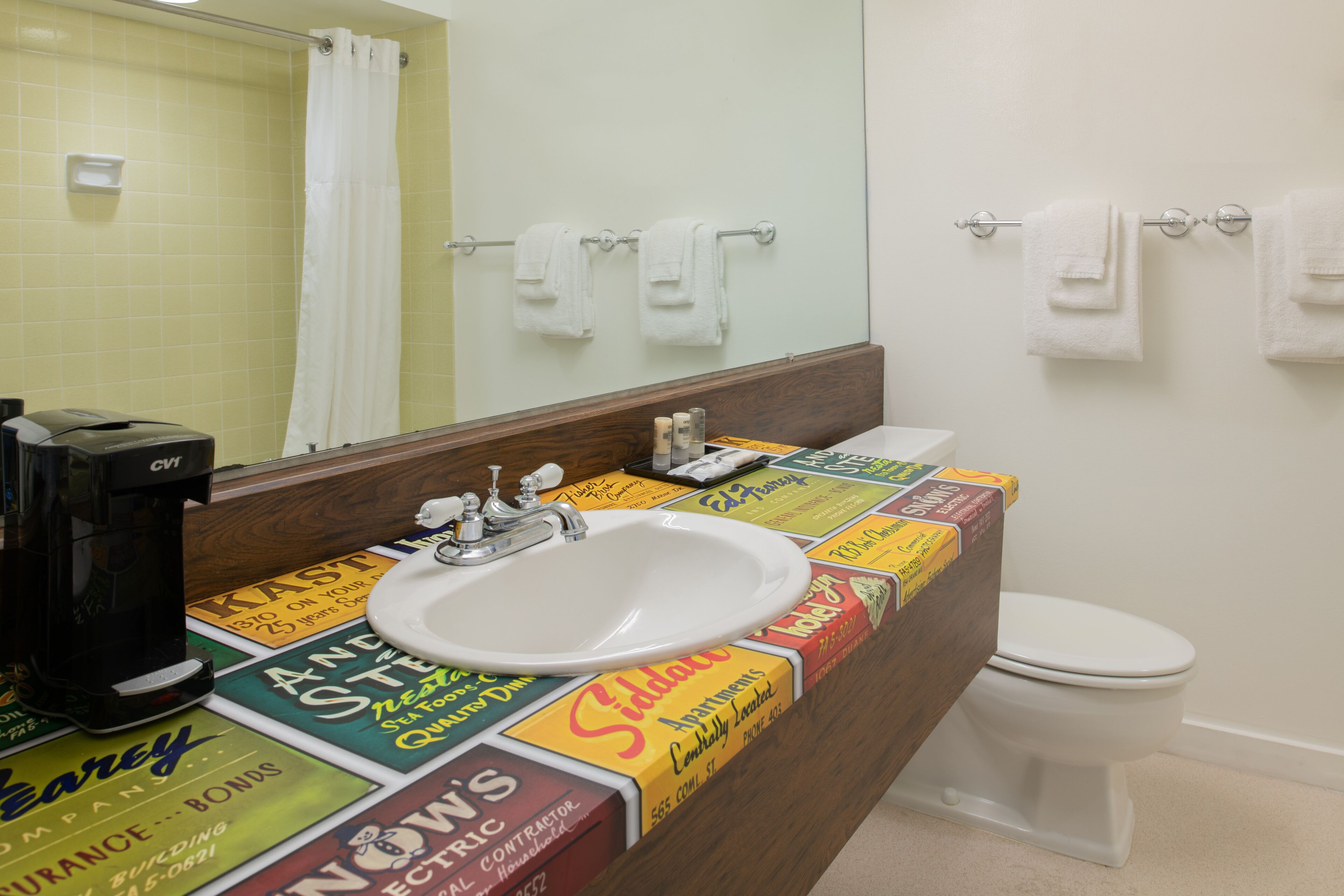 Une salle de bain équipée d’un lavabo, de toilettes et d’un miroir. Le lavabo est posé sur un comptoir où sont disposées diverses publicités.