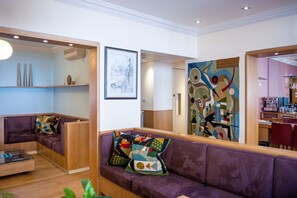 A modern living area with a purple sofa, a glass coffee table, and a wooden bench. There is a large abstract painting on the wall and a framed picture above the sofa. - Sydney House Chelsea (London)