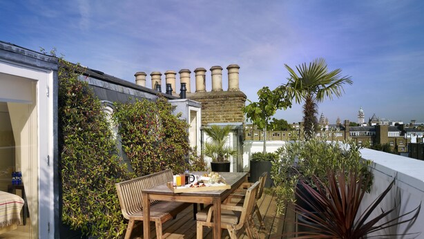 A rooftop terrace with a wooden table and chairs, surrounded by potted plants and a view of historic buildings. - Sydney House Chelsea (London)