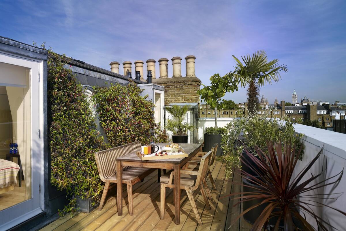 A rooftop terrace with a wooden table and chairs, surrounded by potted plants and a view of historic buildings.