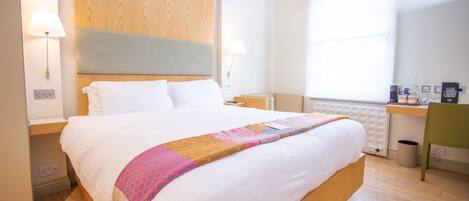 Ein Hotelzimmer mit einem großen Bett, einem Holzkopfteil, Nachttischlampen, einem Schreibtisch mit Stuhl und einem Fenster mit Jalousien.