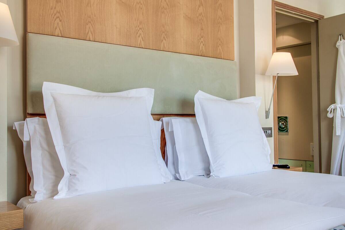 A hotel room with a large bed, wooden headboard, white bedding, a bedside table with a lamp, and a bathroom visible through an open door.