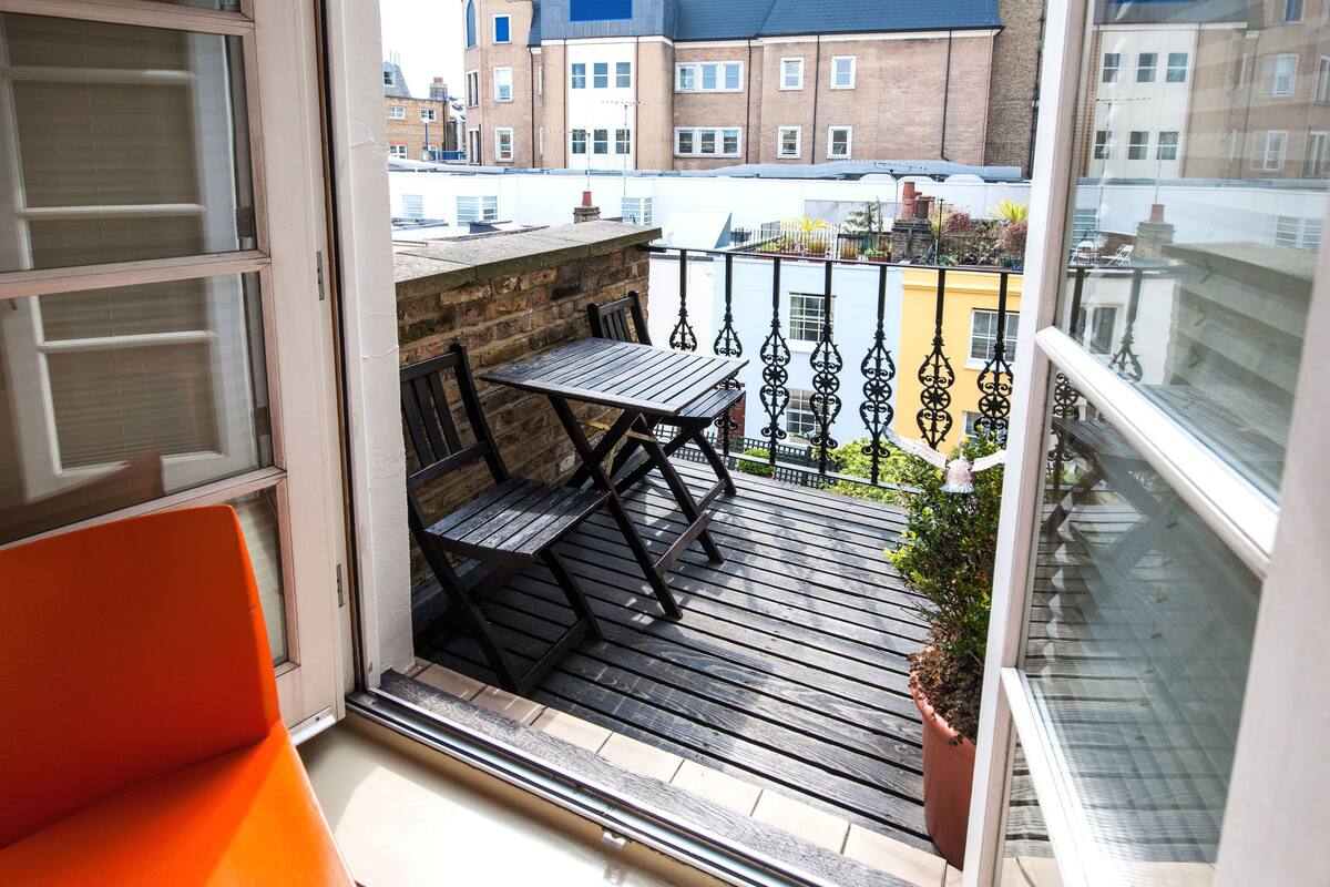 A balcony with a wooden table and chairs, a potted plant, and a view of buildings in the background.