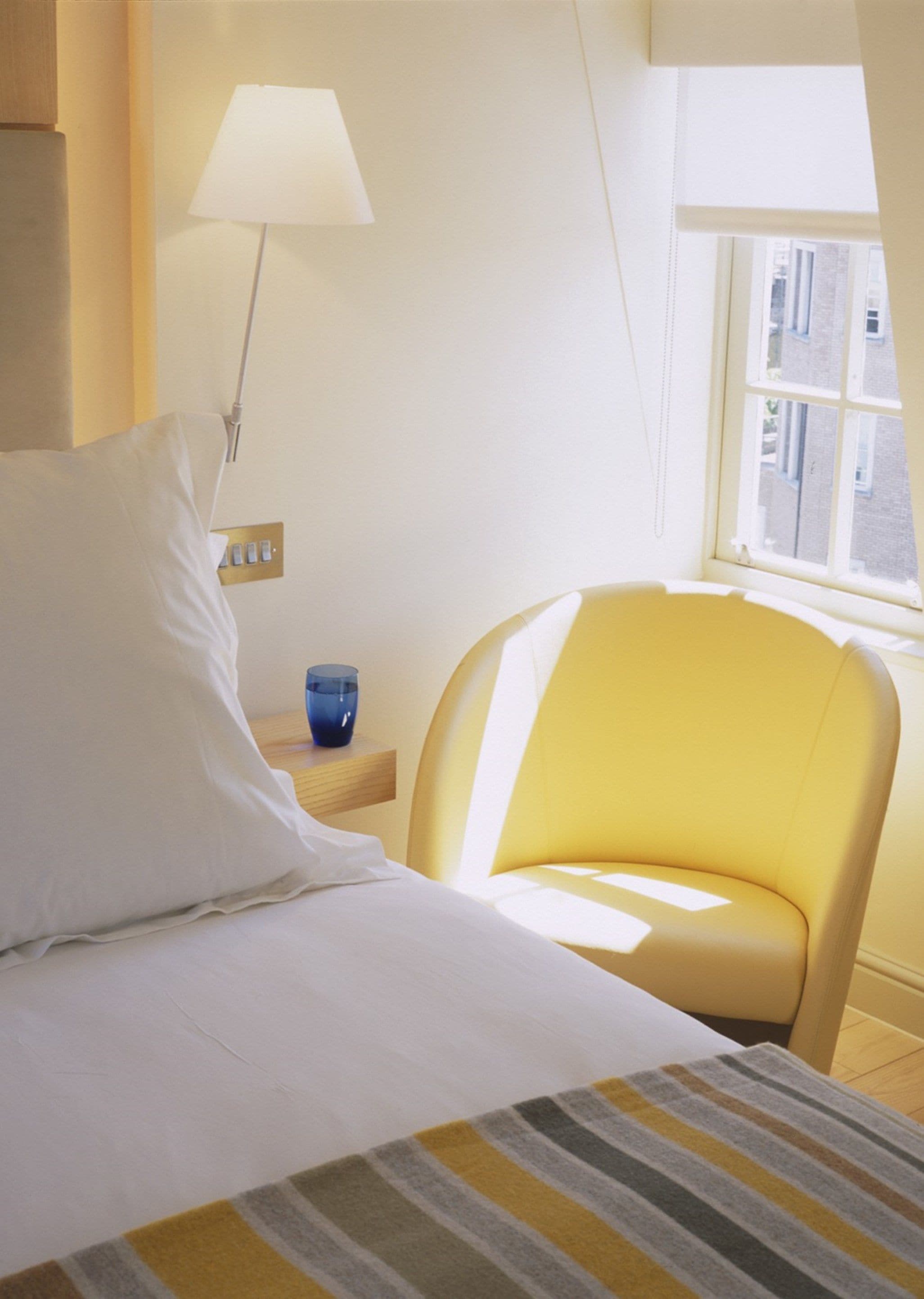 A hotel room with a bed, a nightstand with a lamp, a chair, and a window with a view of buildings.