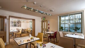 A hotel dining area with multiple tables, chairs, and a large window with plants. - Sydney House Chelsea (London)