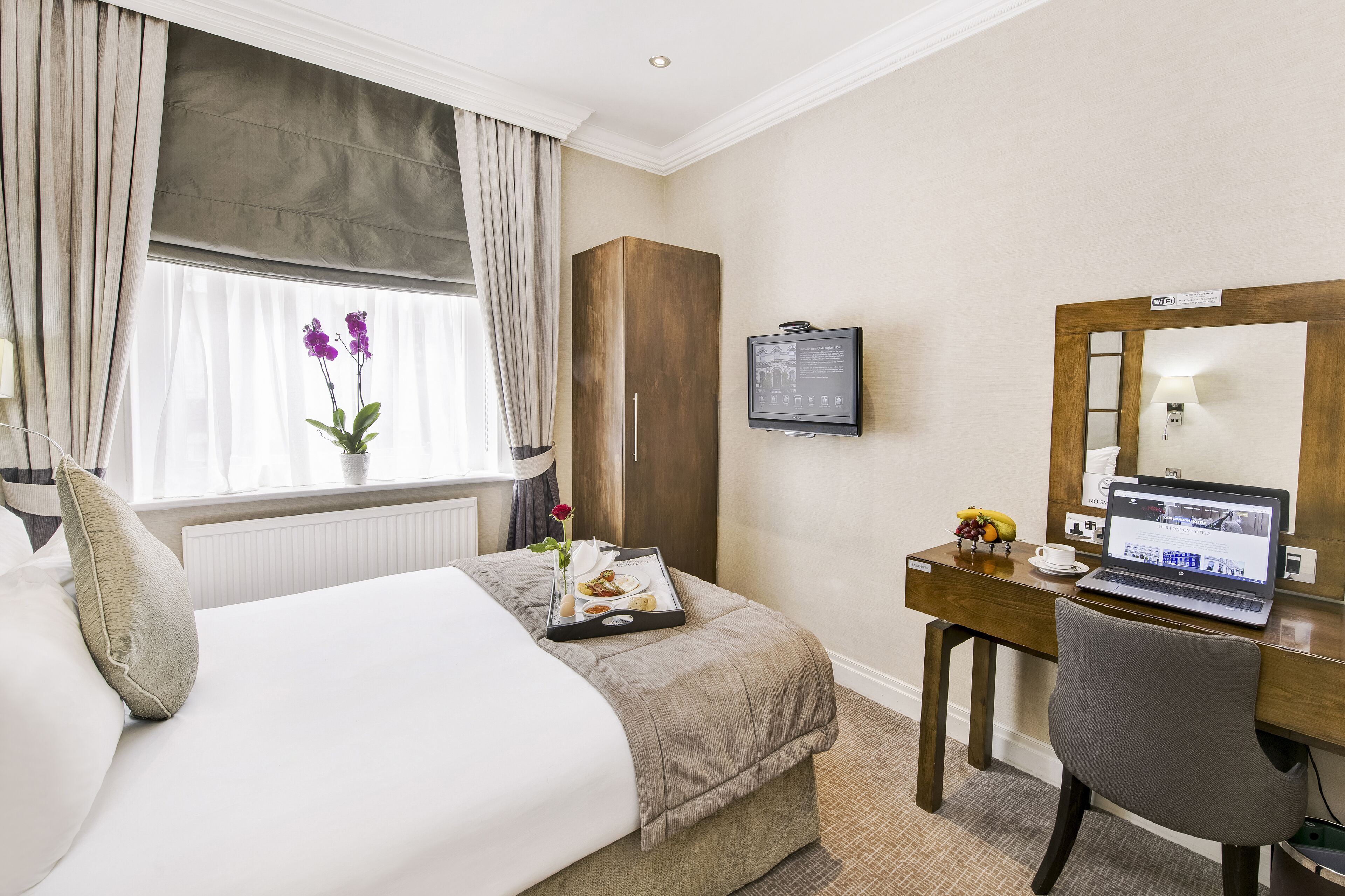 A hotel room with a bed, a desk with a laptop, a TV, and a window with curtains.