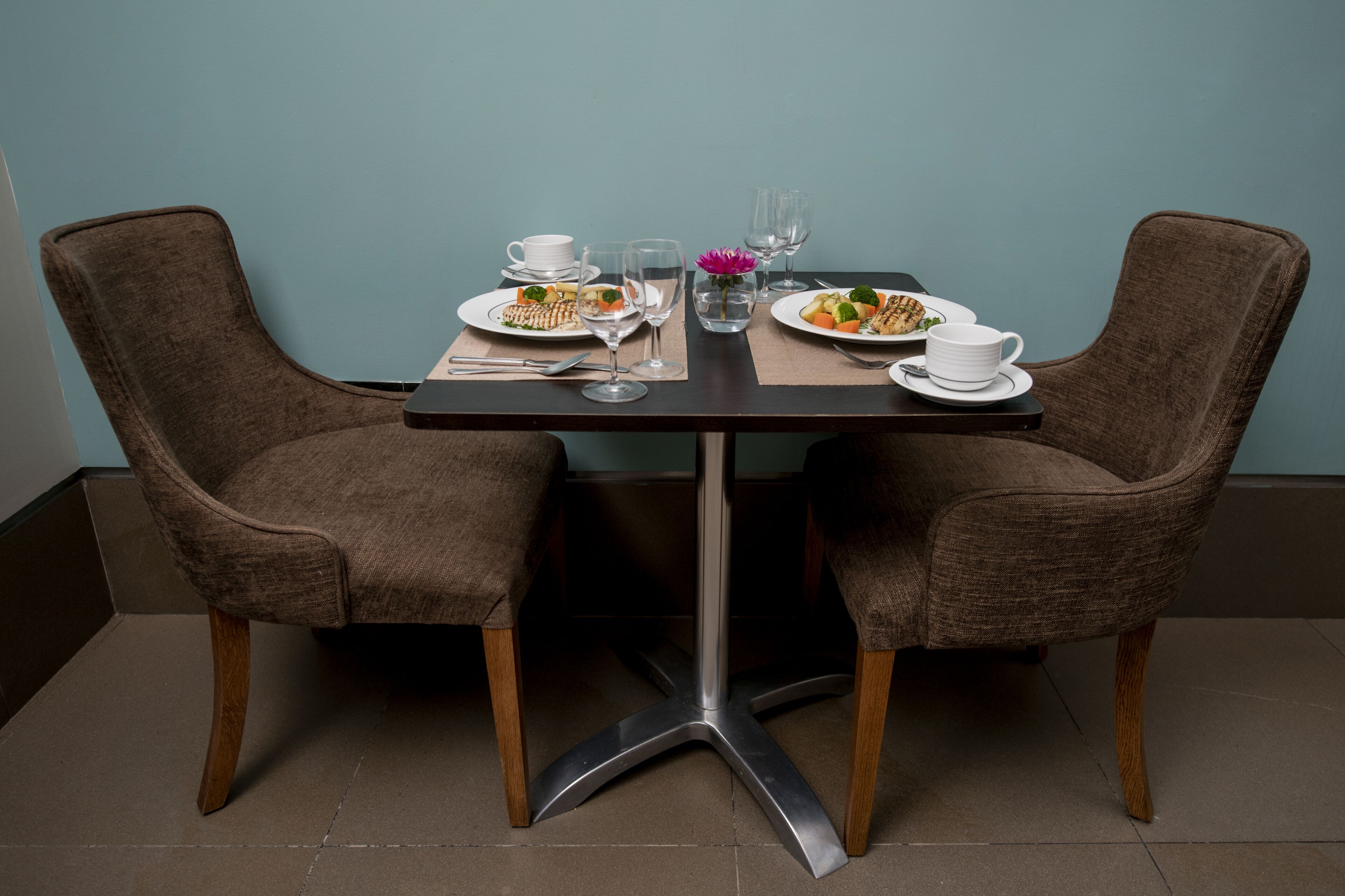 A dining table with two chairs, plates of food, glasses, and a vase with a flower.