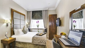 Habitación de hotel con cama, mesita de noche con lámpara, escritorio con una computadora portátil, televisor y ventana con cortinas.