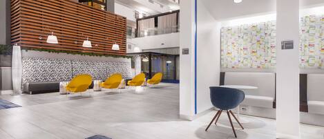 A modern hotel lobby with a wooden slatted wall, yellow chairs, a blue chair, and a patterned wall.