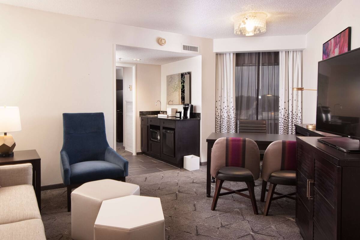 A hotel room with a seating area, a TV, a kitchenette, and a large window with curtains.