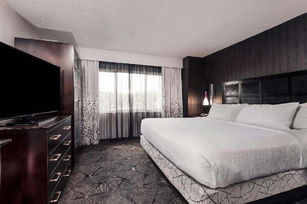 A hotel room with a large bed, a TV, a dresser, and a window with curtains.