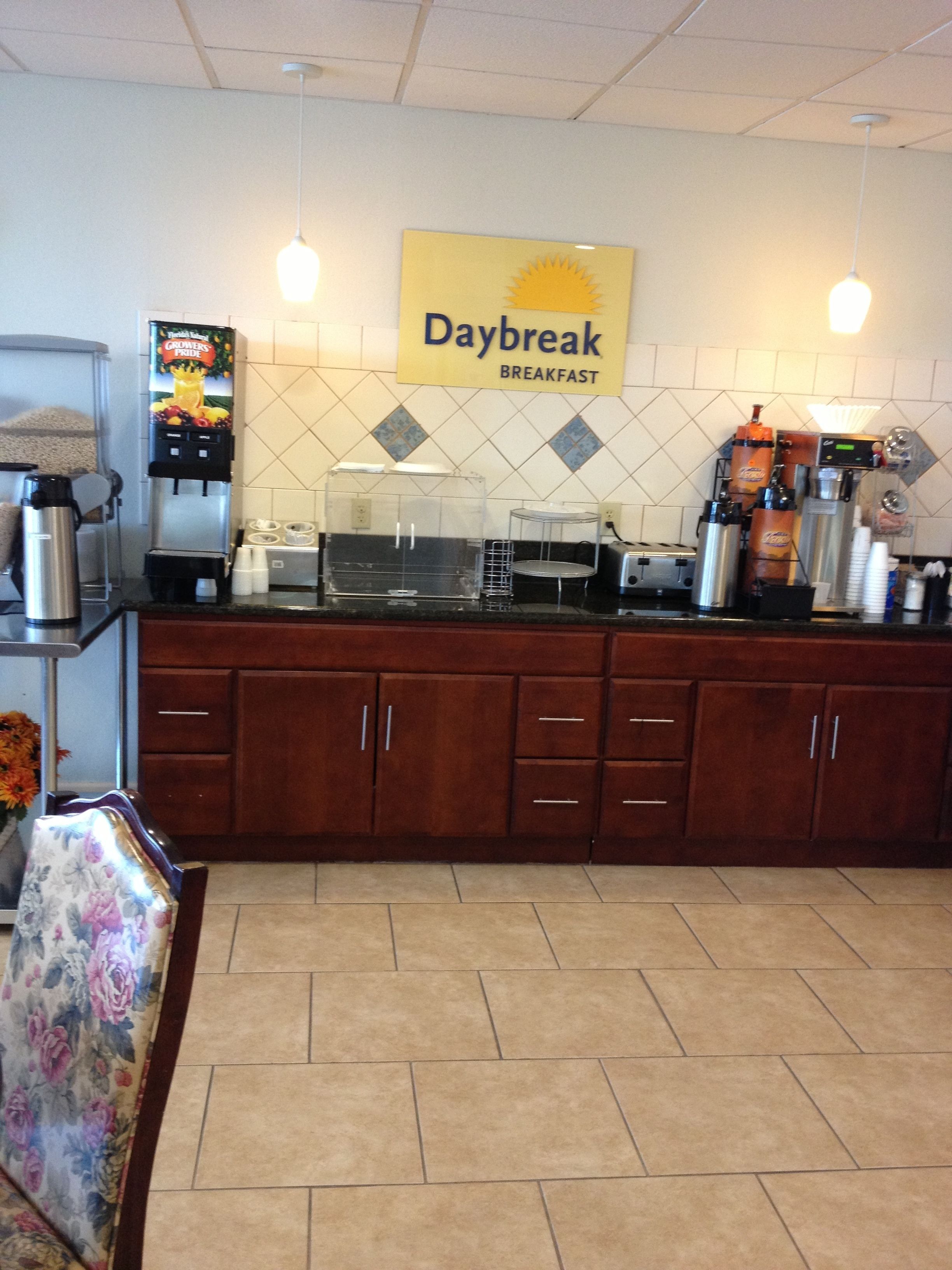 Un área de desayuno con una máquina de café, un dispensador de zumo y un cartel que dice "Daybreak Breakfast".