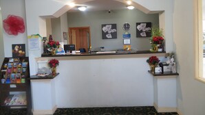 Zona de recepción con escritorio, computadora y obras de arte enmarcadas. Hay arreglos florales y un reloj en la pared.