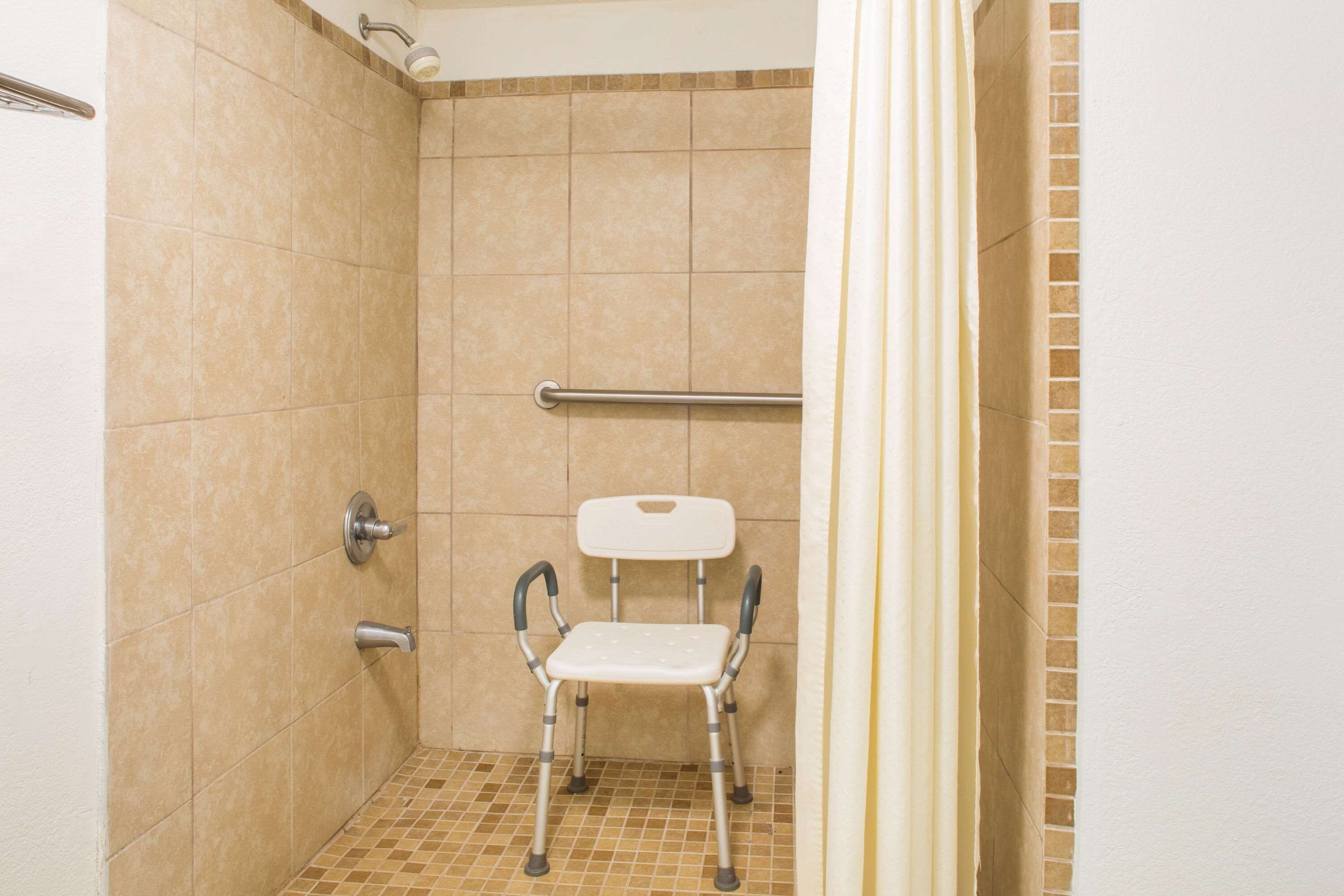 Un espace douche avec des carreaux de couleur beige, une chaise blanche et une barre d’appui.
