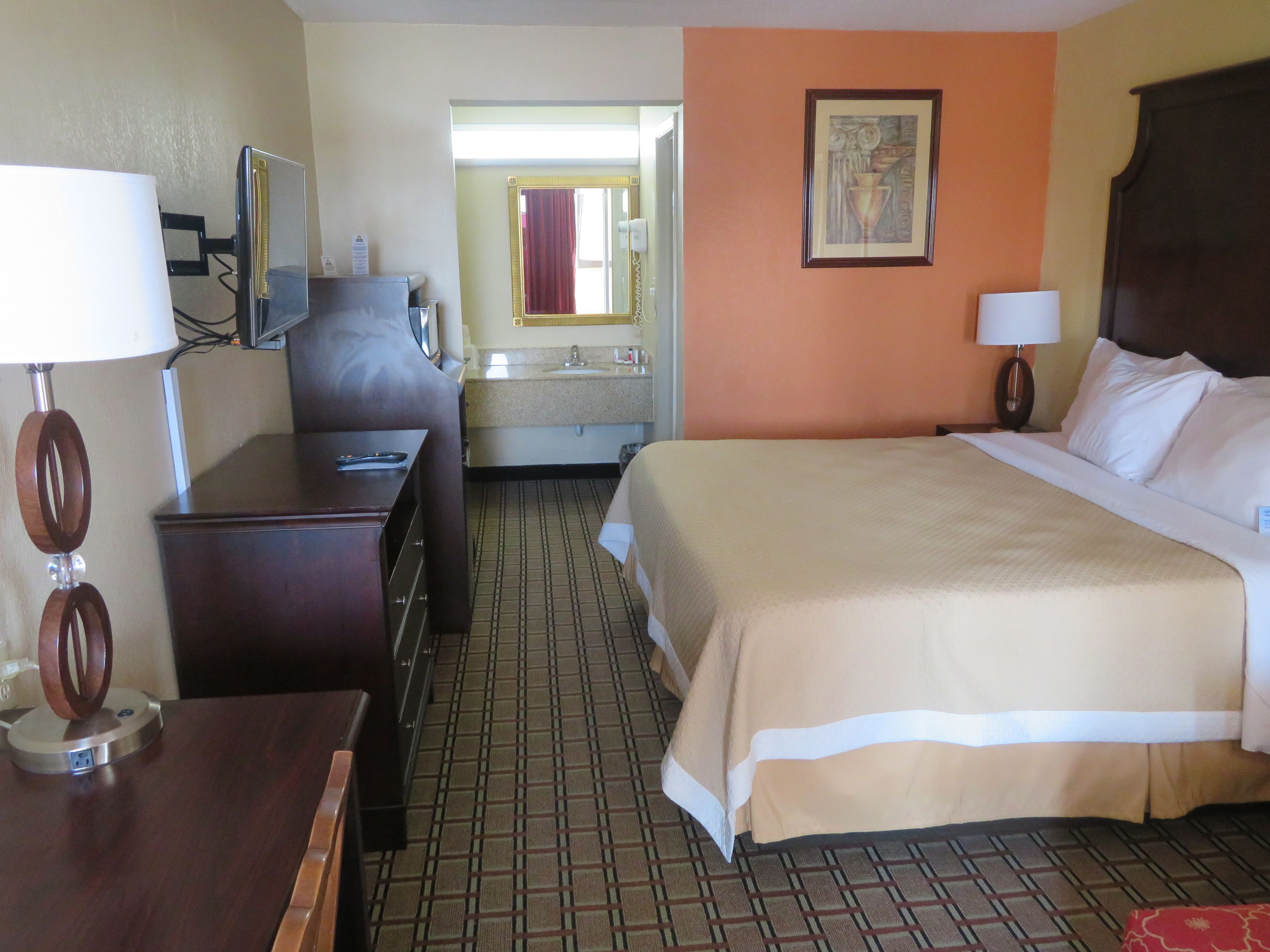 A hotel room with a bed, bedside table, desk, chair, and a bathroom visible through an open door.