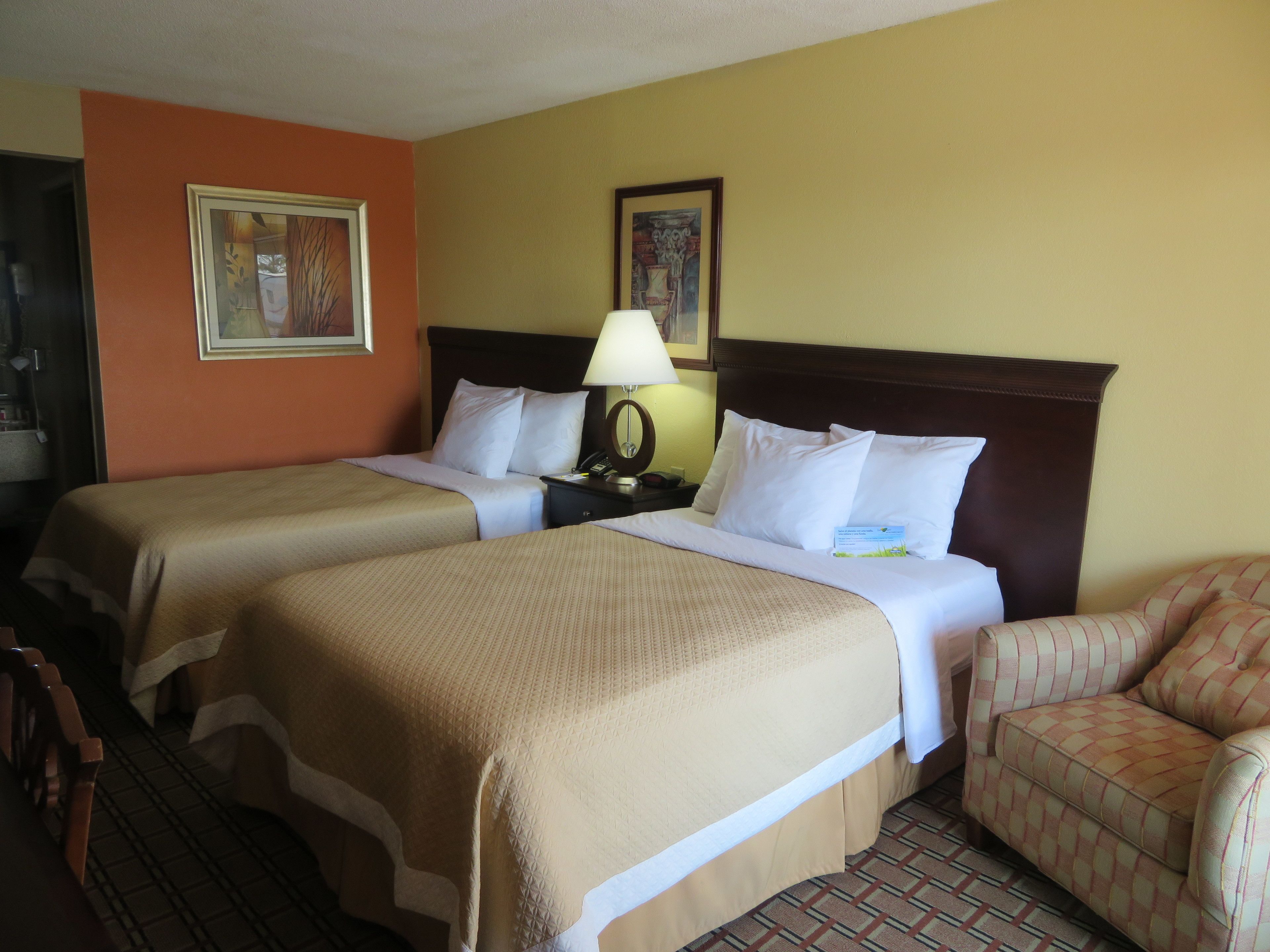 A hotel room with two beds, a nightstand with a lamp, a chair, and two framed pictures on the wall.