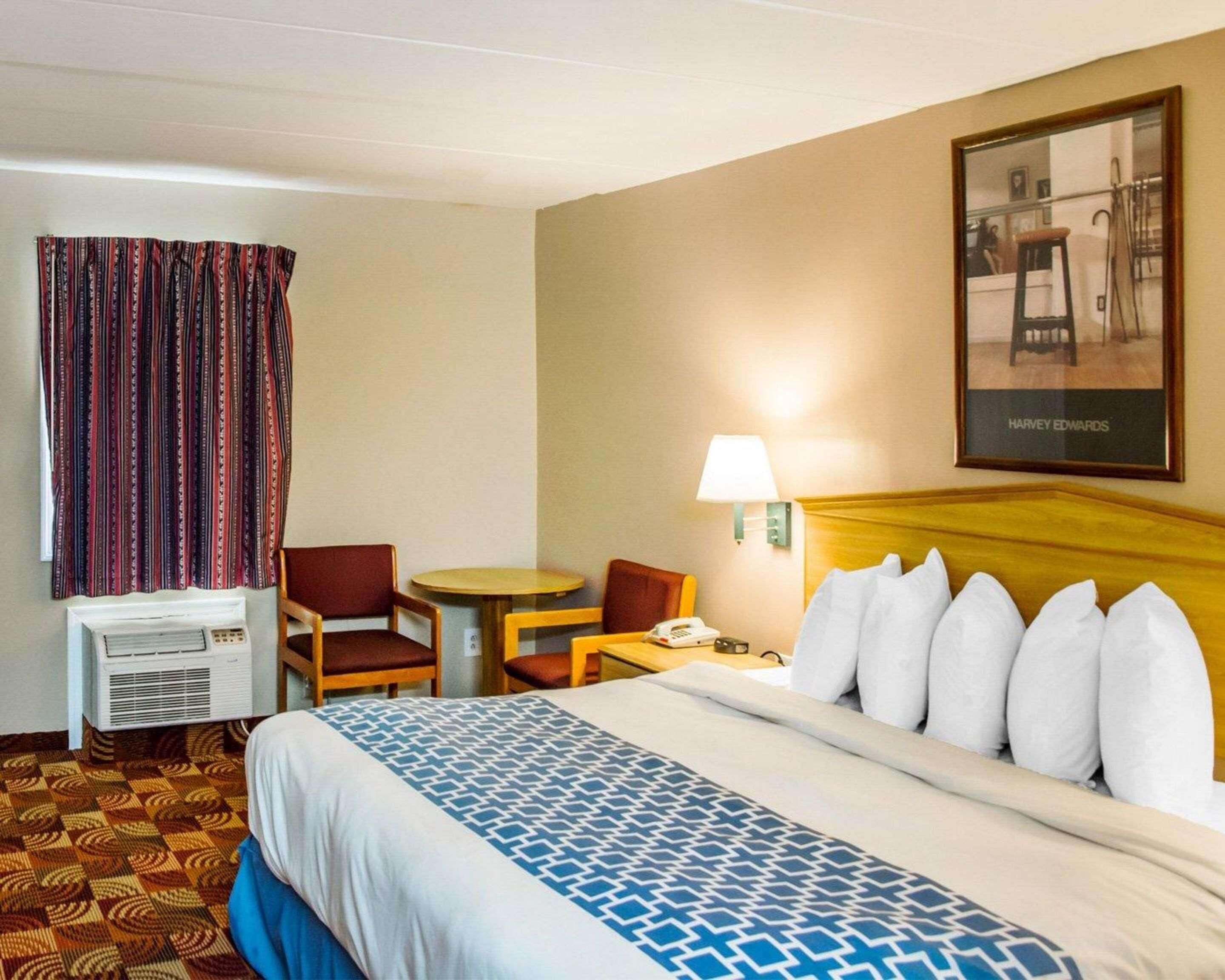A hotel room with a bed, a desk, a chair, a lamp, a picture frame, and an air conditioning unit.