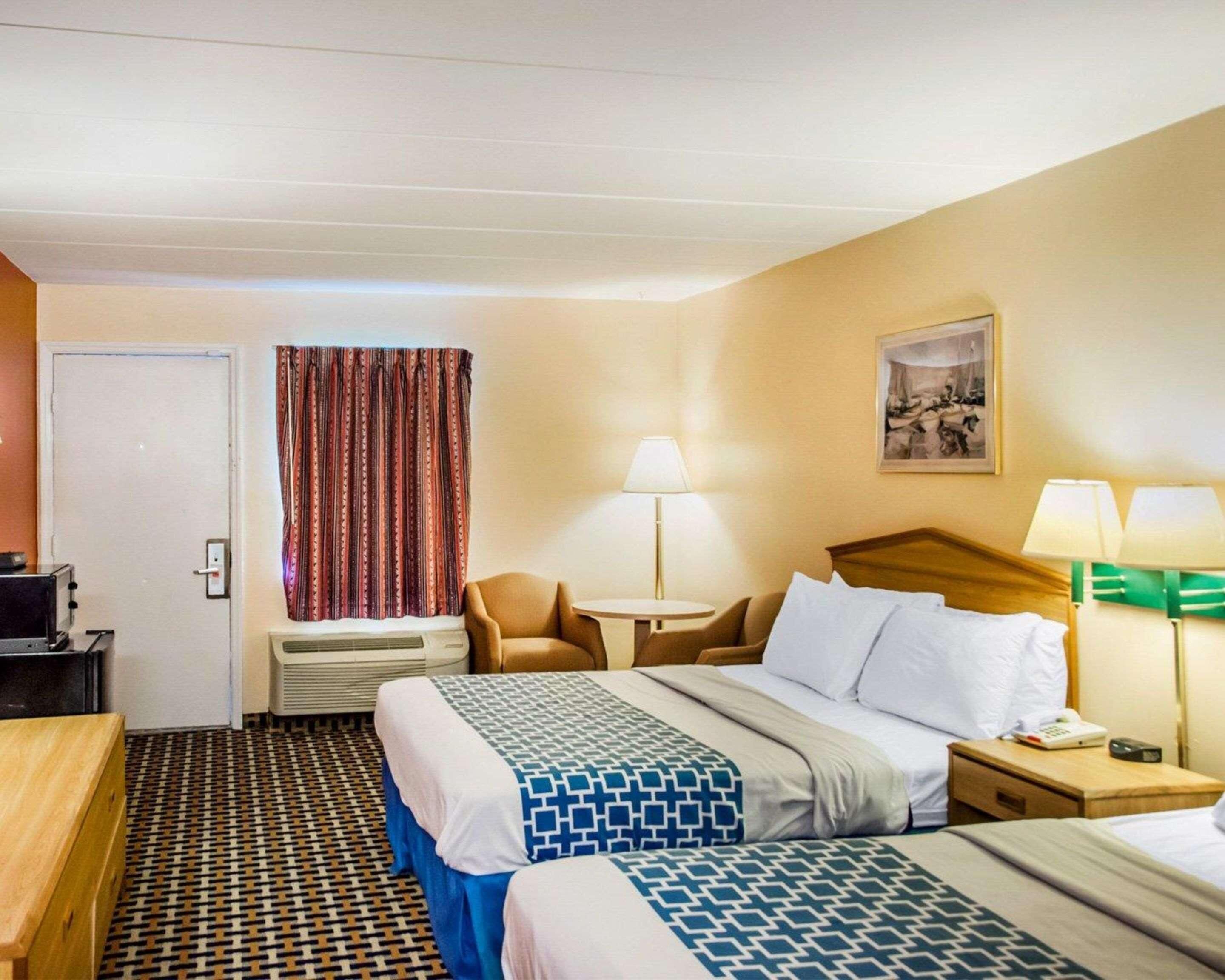 A hotel room with a bed, two bedside tables, a chair, a desk, a lamp, a painting, and a patterned floor.