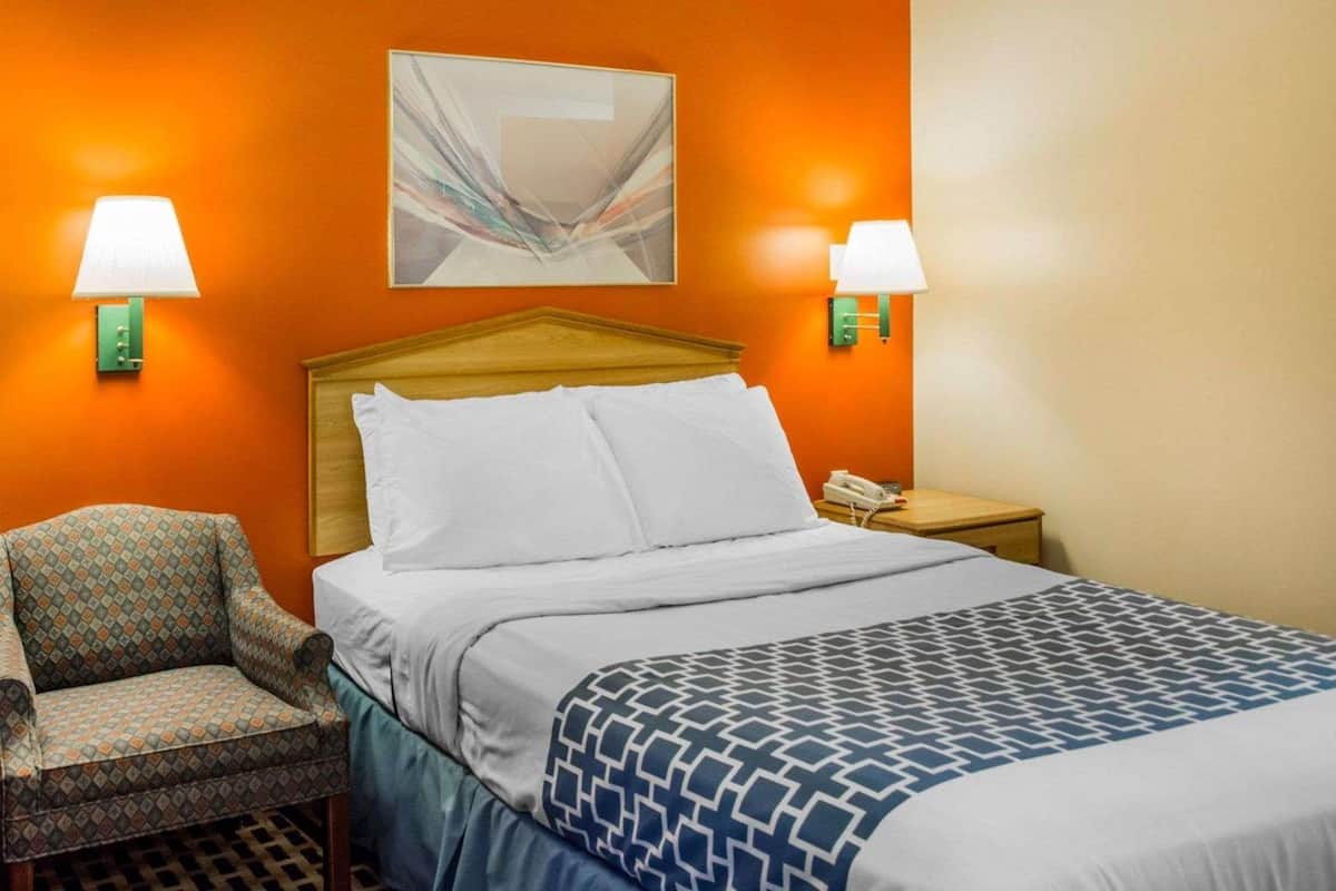 A hotel room with a bed, two chairs, a nightstand with a phone, and wall-mounted lamps.