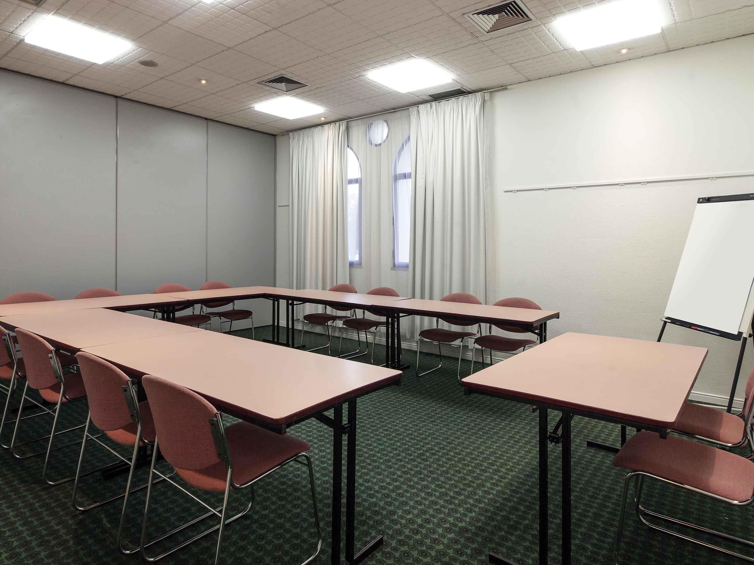 Ein Konferenzraum mit einem langen Tisch, Stühlen, einer Whiteboard-Tafel und Fenstern mit Vorhängen.