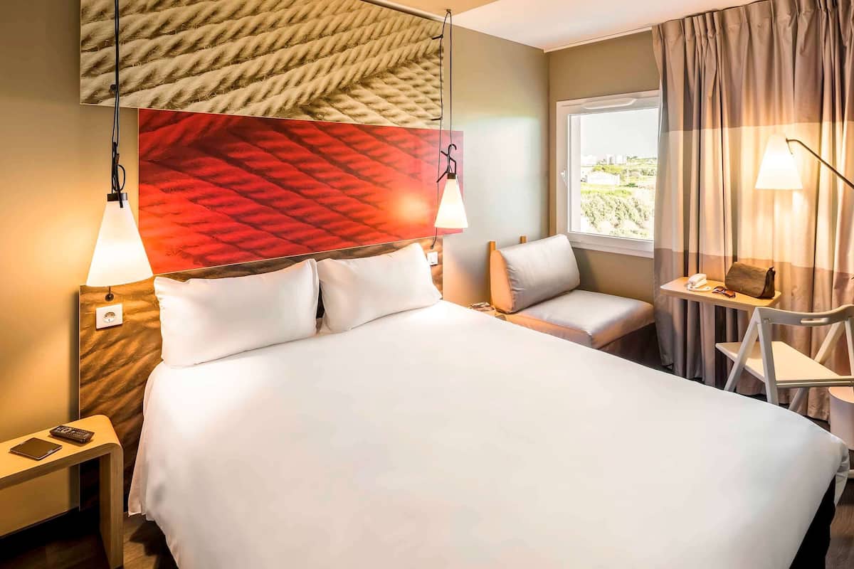 Ein Hotelzimmer mit einem großen Bett, Nachttischlampen, einem kleinen Tisch mit Telefon und Gläsern, einem Sessel und Blick auf Grünflächen.