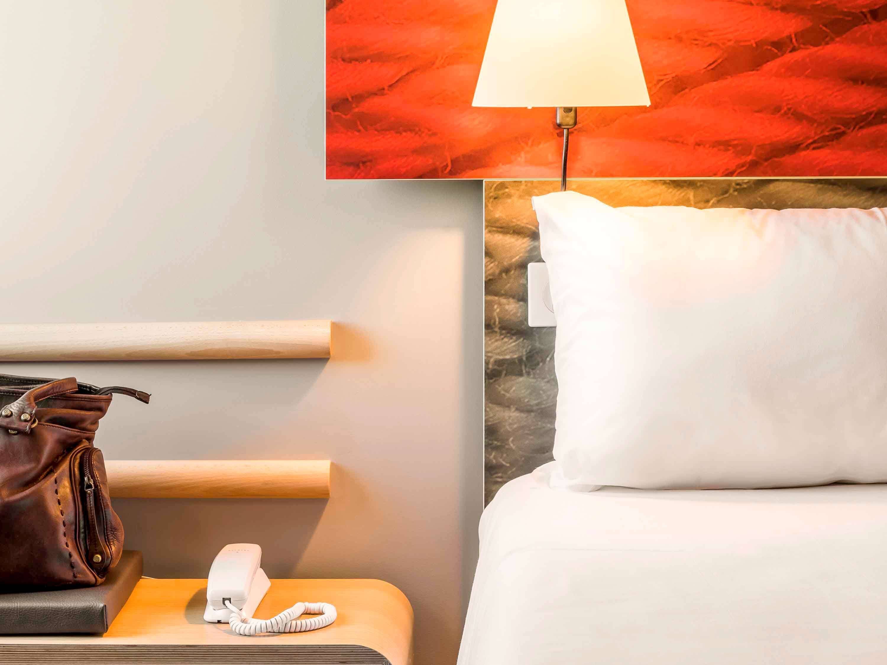 A hotel room with a bed, a nightstand with a lamp, a telephone, and a brown leather bag.