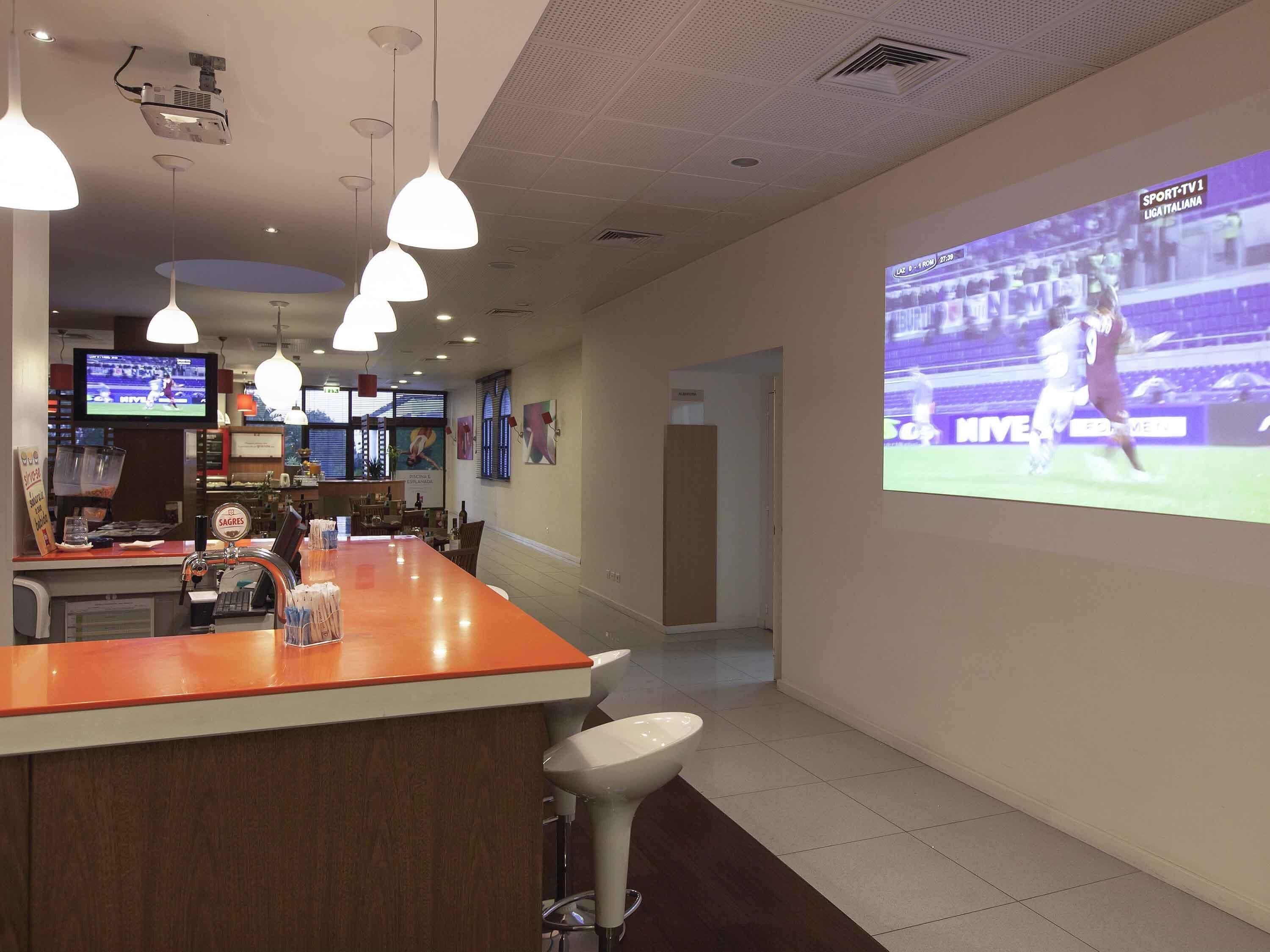 A bar area with a projection screen displaying a sports event, a counter with a coffee machine, and hanging pendant lights.