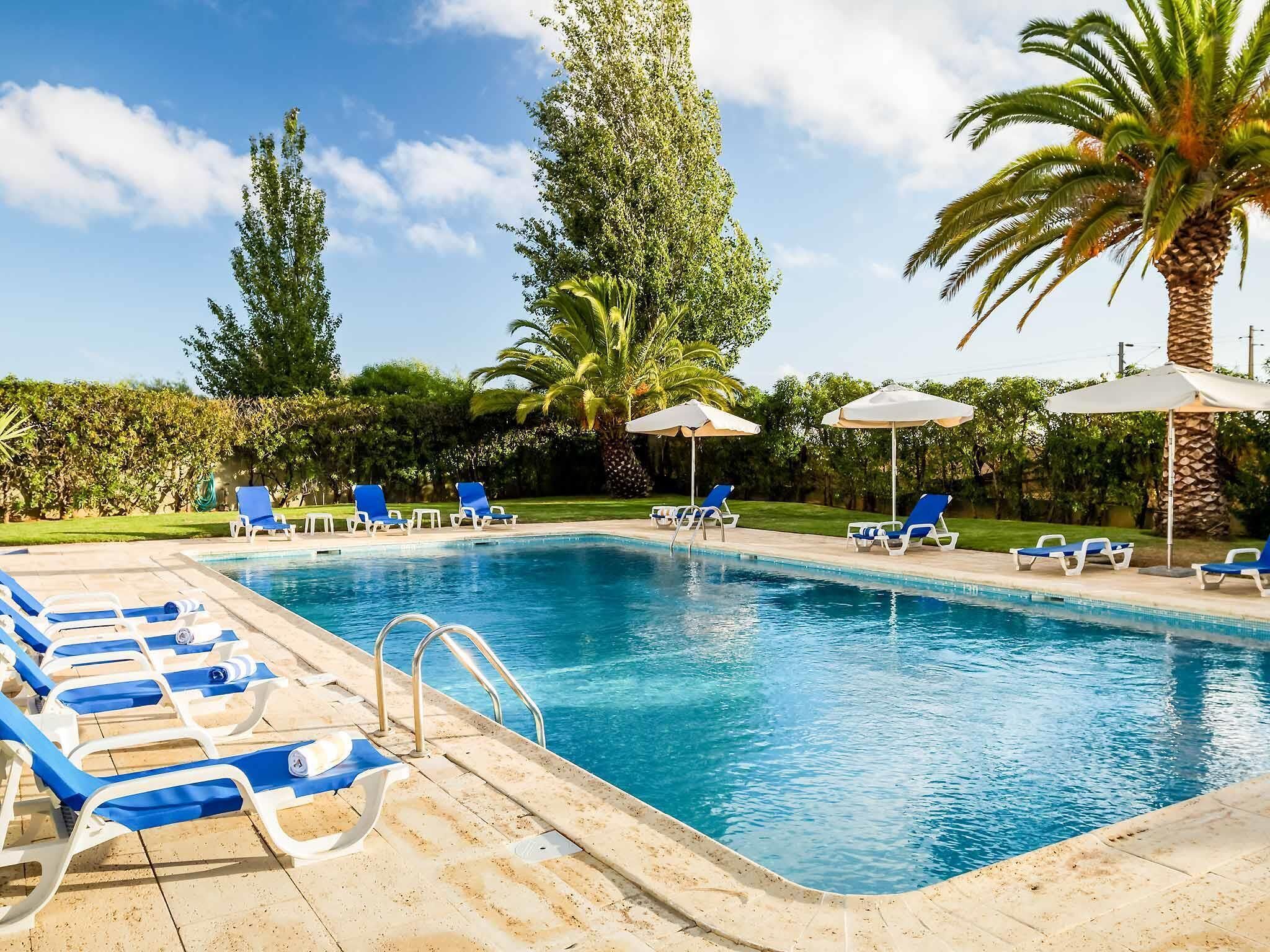 Piscina con sillas de descanso azules y sombrillas, rodeada de palmeras y follaje.