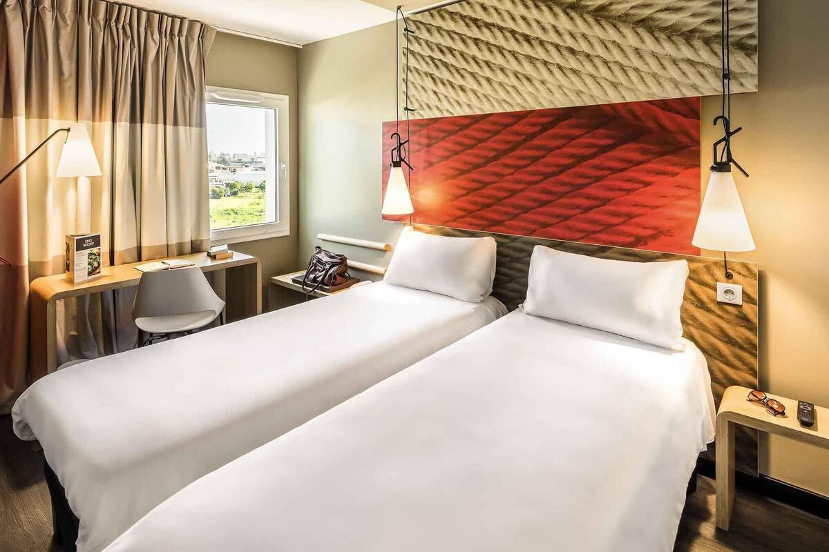 Ein Hotelzimmer mit zwei Betten, einem Schreibtisch, einem Stuhl, einem Fenster mit Ausblick und einer roten Wand mit Struktur.