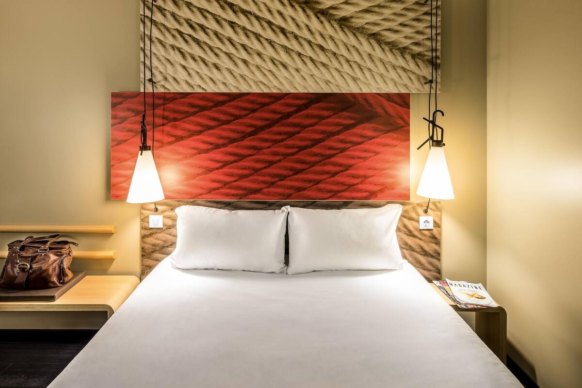 Ein Hotelzimmer mit einem Bett, zwei Nachttischlampen, einem Nachttisch mit einer Handtasche und einer Zeitschrift auf dem Nachttisch.