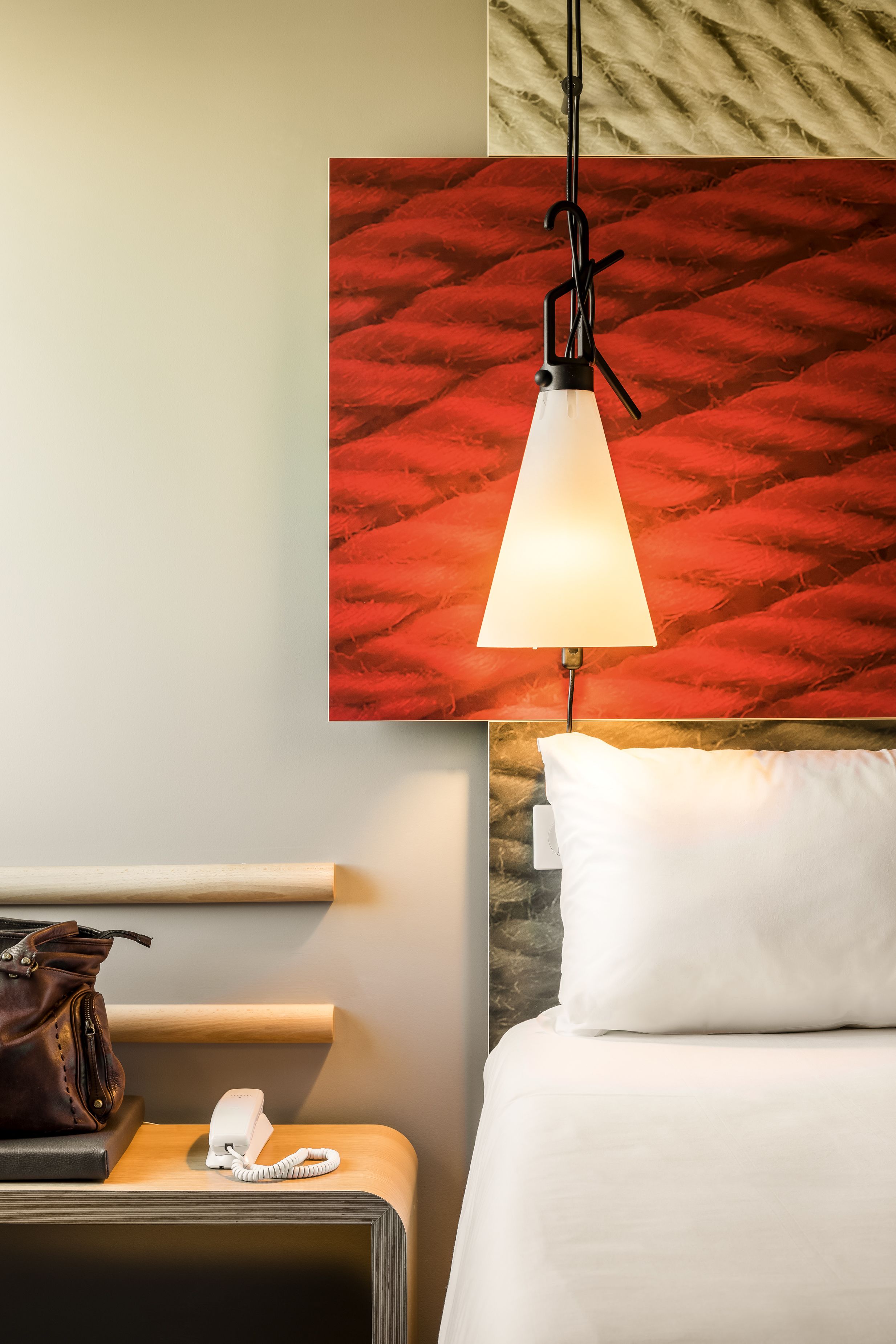 A hotel room with a bed, a bedside table with a phone, a lamp, and a red textured wall.