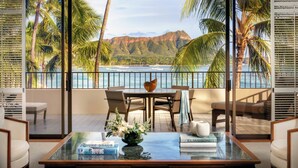 A modern living room with a glass coffee table, books, and a decorative bowl. It features a view of a beach and mountains through large windows. - Halekulani (Honolulu)