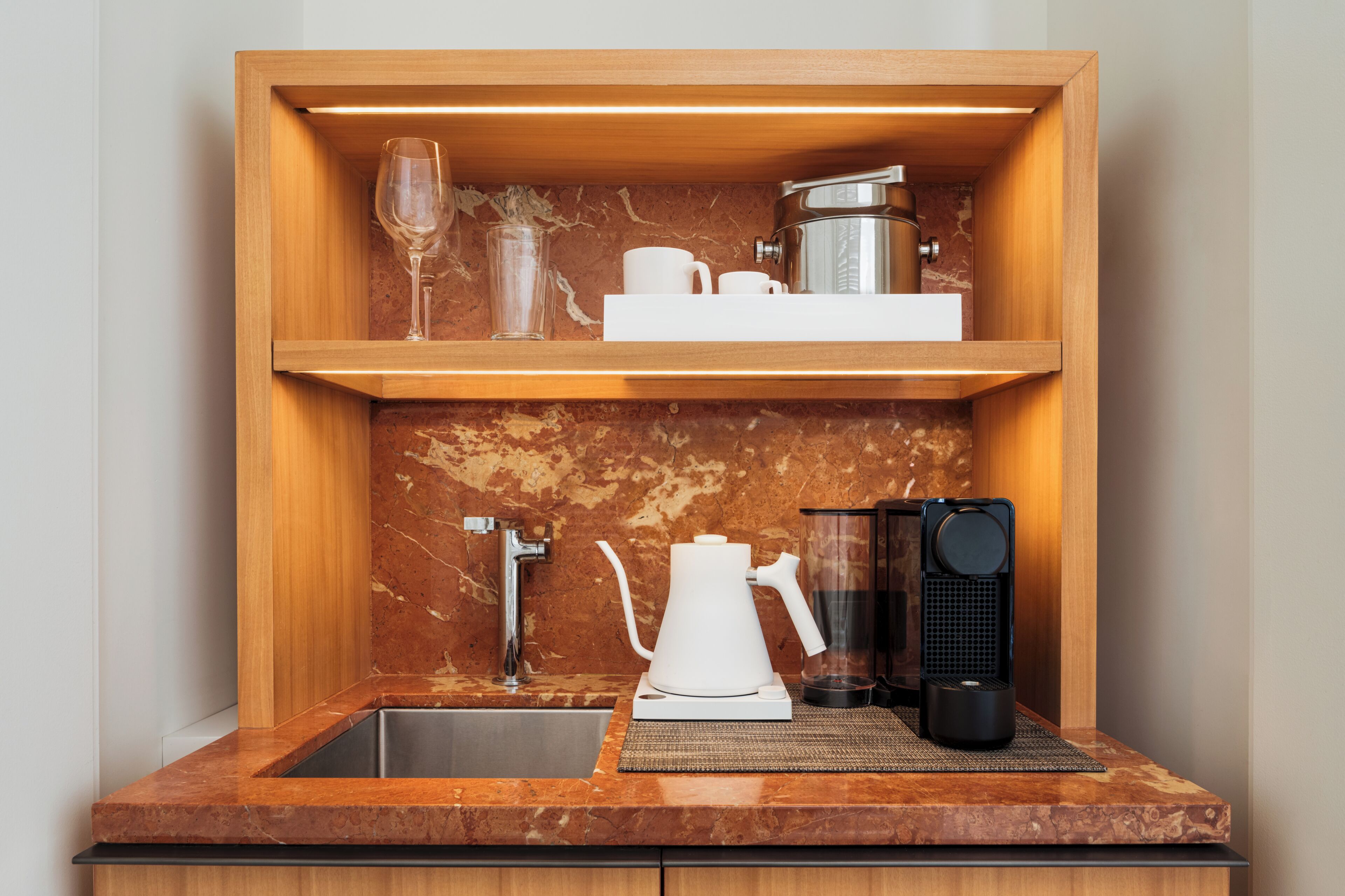 Une cuisine compacte dotée d’un plan de travail en marbre, d’un évier, d’une machine à café, d’une bouilloire et d’une étagère avec un verre et des tasses.